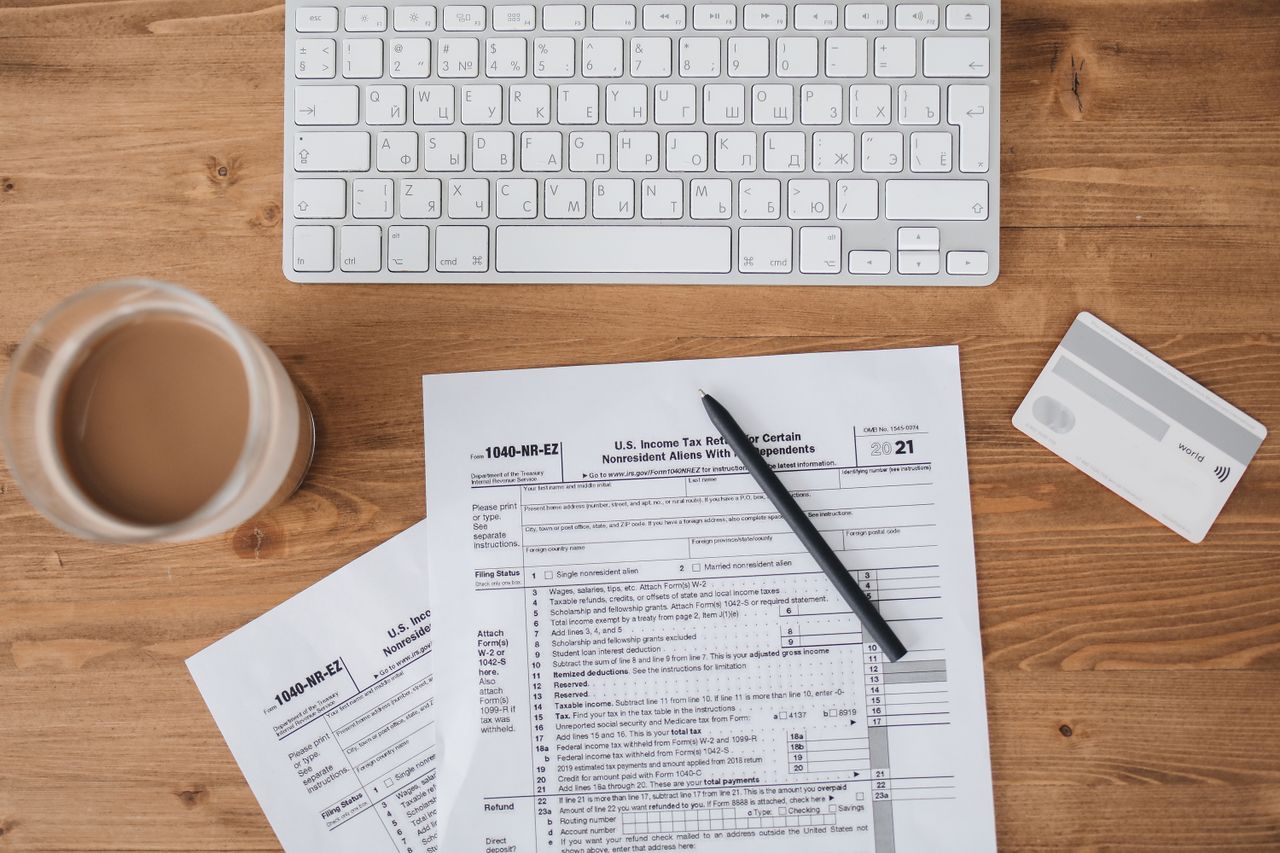 cta-bg Top view of tax forms, keyboard, pen, and coffee on a wooden desk.